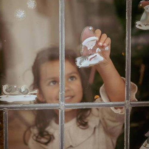 Bambina posiziona adesivi natalizi di Little Dutch su una finestra, inclusi un albero di Natale, fiocchi di neve e figure festose.