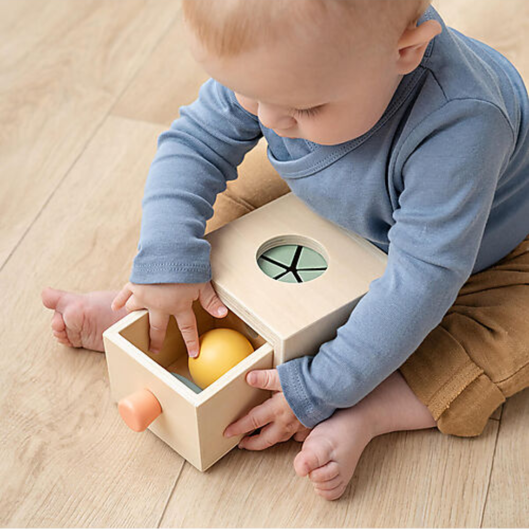 Bambino che interagisce con il cassetto del Gioco Montessori Permanence Box in Legno, recuperando la pallina gialla e sviluppando la motricità fine.