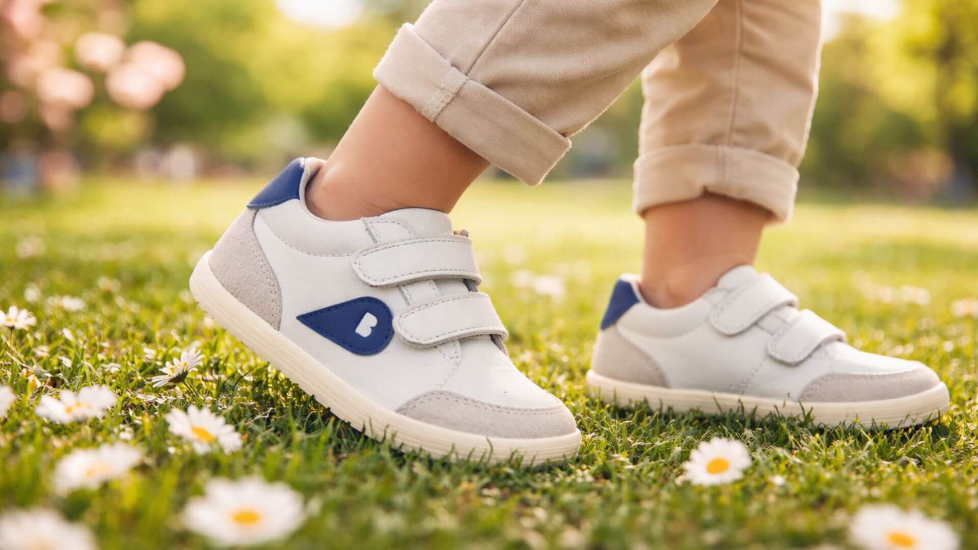 Bambino che cammina in un prato con scarpe primi passi Bobux bianche e blu, calzature barefoot e morbide ideali per lo sviluppo naturale del piede