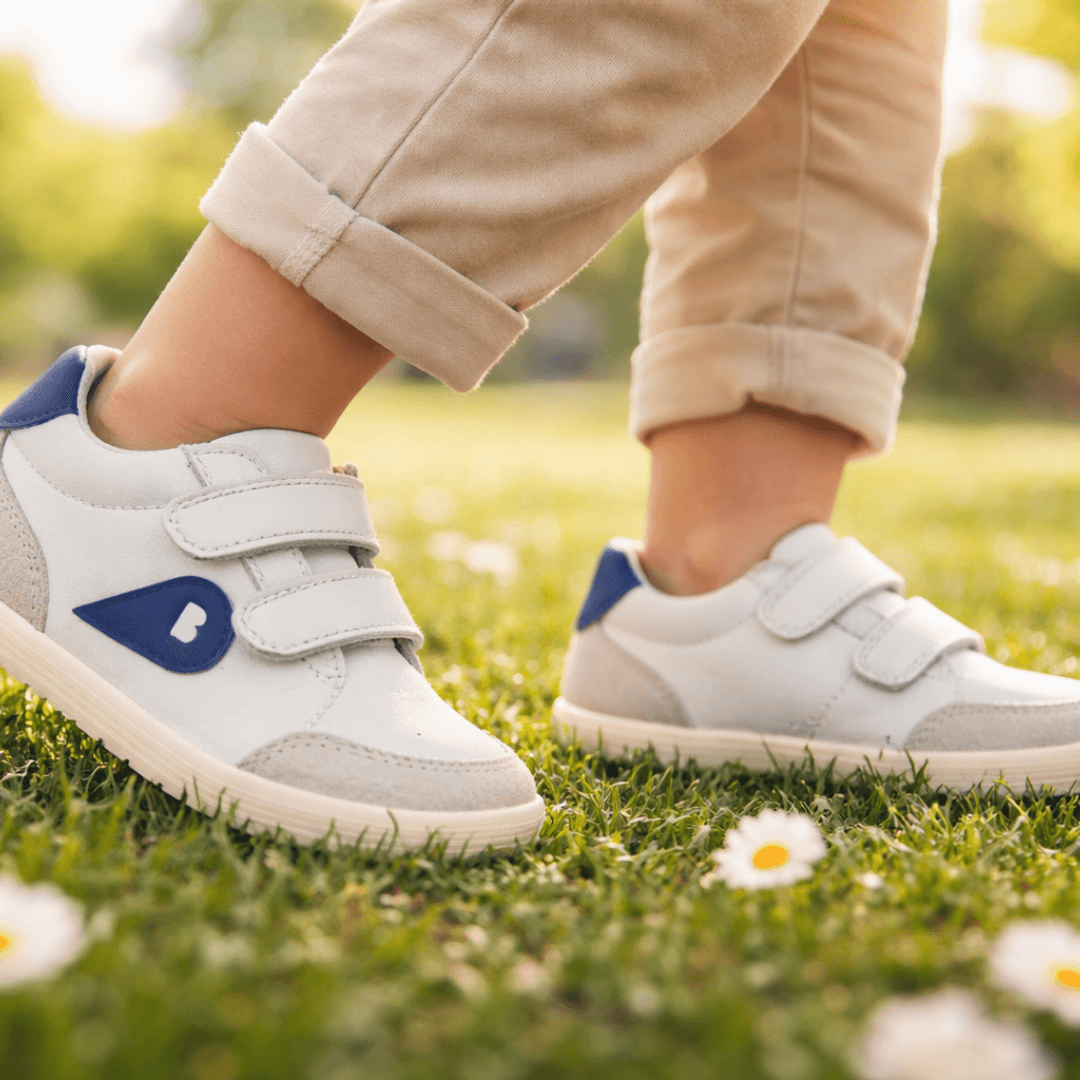 Bambino che cammina in un prato con scarpe primi passi Bobux bianche e blu, calzature barefoot e morbide ideali per lo sviluppo naturale del piede