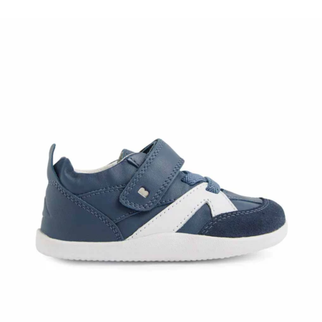 Un paio di scarpe primi passi morbide Bobux Xplorer Chase colore Vintage Indigo in pelle blu con lacci elastici e chiusura a strappo, appoggiate su un pavimento in legno chiaro e illuminate dalla luce naturale.